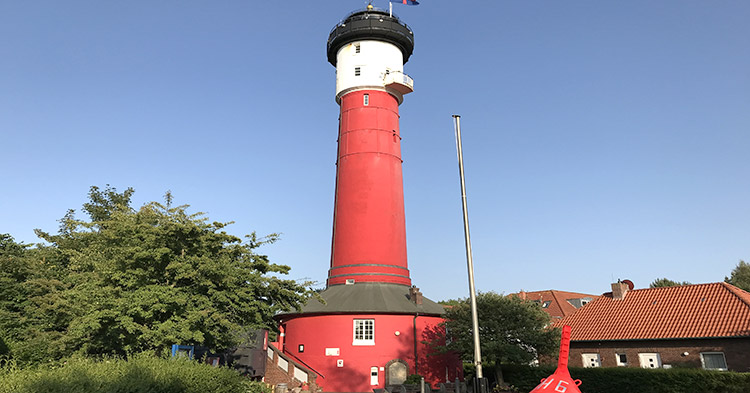 Der alte rot-weiße Leuchtturm von Wangerooge ragt vor blauem Himmel in die Höhe, umgeben von Bäumen und historischen Backsteingebäuden