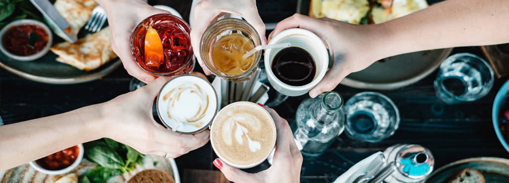 Hände fünf verschiedener Personen von oben, die über einem gedeckten Tisch mit Kaffee, Cocktails und Softdrinks anstoßen