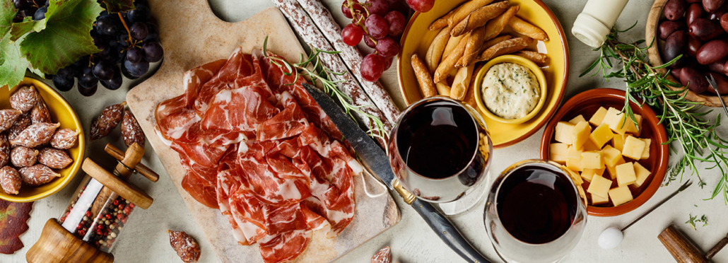 Auswahl verschiedener Tapas, bestehend aus Schinken, Salami, Käsewürfeln, Oliven und Brotstangen, dazwischen zwei Gläser Rotwein
