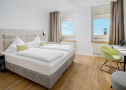 Helles Schlafzimmer einer Suite im Parkhotel Wangerooge mit großem gemachtem Doppelbett, Sessel, Schreibtisch und Leuchtturmblick