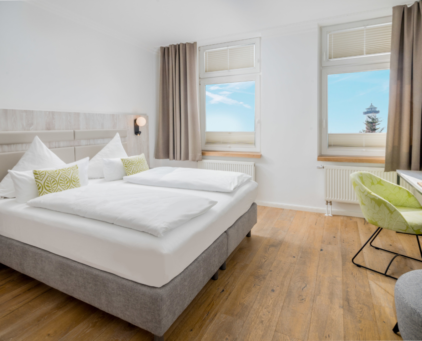 Helles Schlafzimmer einer Suite im Parkhotel Wangerooge mit großem gemachtem Doppelbett, Sessel, Schreibtisch und Leuchtturmblick