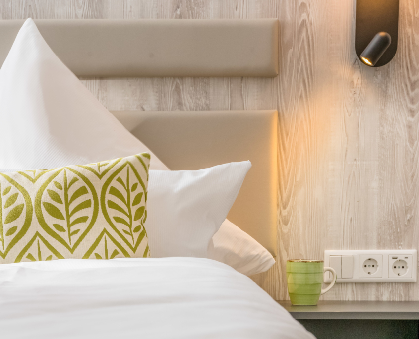 Detailaufnahme eines Doppelbetts im Parkhotel Wangerooge mit Kopfteil in Holzoptik, weißer Bettwäsche, grün gemustertem Zierkissen und eingeschalteter Wandlampe
