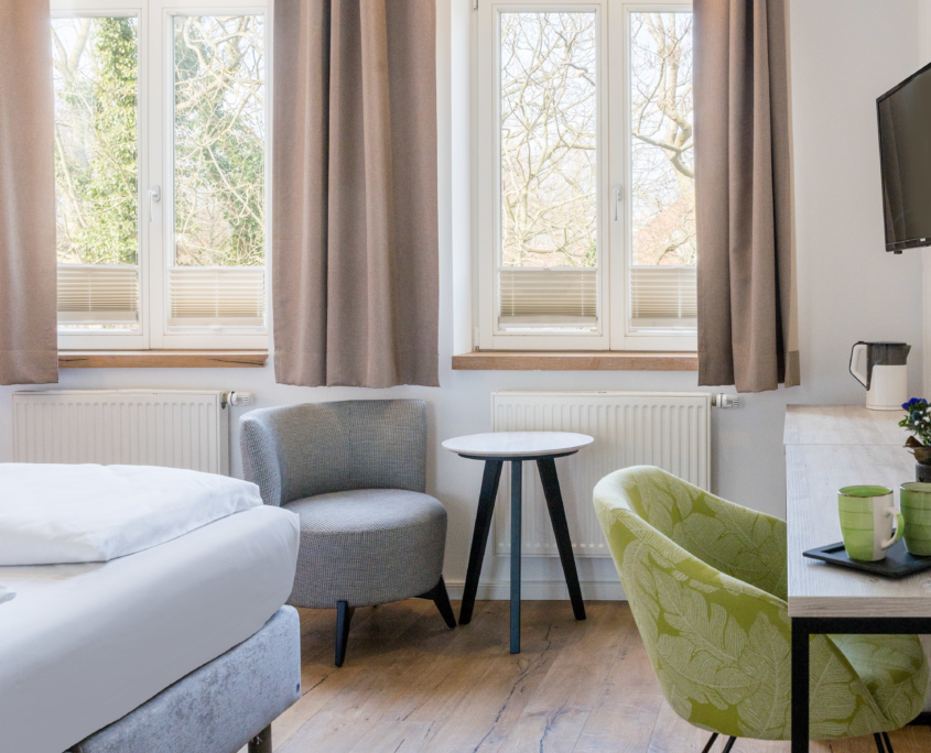 Nahaufnahme von Sitzbereich und Schreibtisch in einem geräumigen Doppelzimmer des Parkhotel Wangerooge mit großen Doppelfenstern und Blick ins Grüne