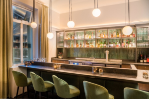 Moderne Hausbar im Parkhotel Wangerooge mit kugelförmigen Hängelampen über dem Tresen, Barhockern aus grünem Samt und großer Spirituosenauswahl im beleuchteten Barregal an der Wand