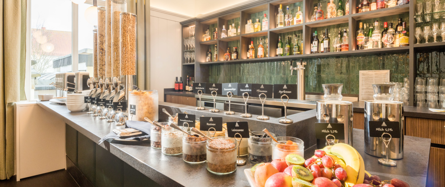Frühstückstresen im Parkhotel Wangerooge mit vielfältiger Müsliauswahl, frischem Obst, Nüssen, Kernen, Trockenfrüchten und Milch vor einem modernen Barregal